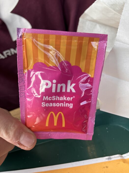 En påse med "Pink McShaker Seasoning" från McDonald's håller upp av en hand, mot en bakgrund av tyg och papper.