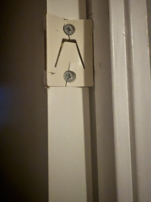 Närbild av en dörrkarm med en vit plastdel fastsatt med två skruvar, eventuellt en del av ett lås- eller stängningsmekanism.