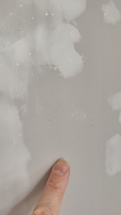 Finger pekar på små gropar samt ojämnheter på en bredspacklad vägg, med ljusa fläckar som kan vara spackelrester eller slipmärken.