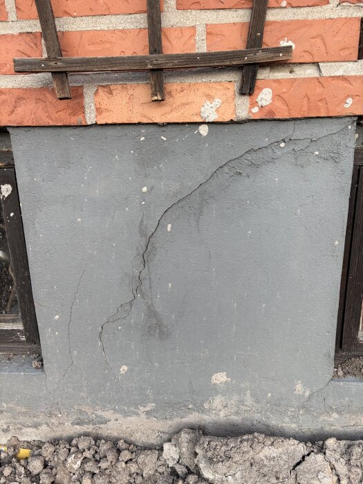Spricka i grå cementfasad under röda tegelstenar. Seen under en trästruktur, marken nedanför visar lös jord och stenar, vilket indikerar potentiell skada.