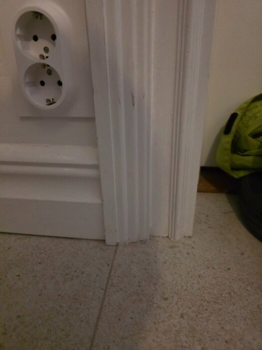 Närbild av listning och panel i kök med nybrofoder, eluttag syns på väggen ovan. Golv med ljusbrun platta.