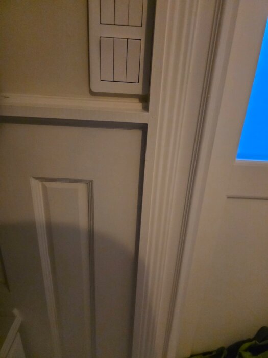 Vägg med panel, lister och dörrkarm i ett rum. Syns en ljusströmbrytare och skuggor på panelen. Renoveringsdetaljer visas i en hall.