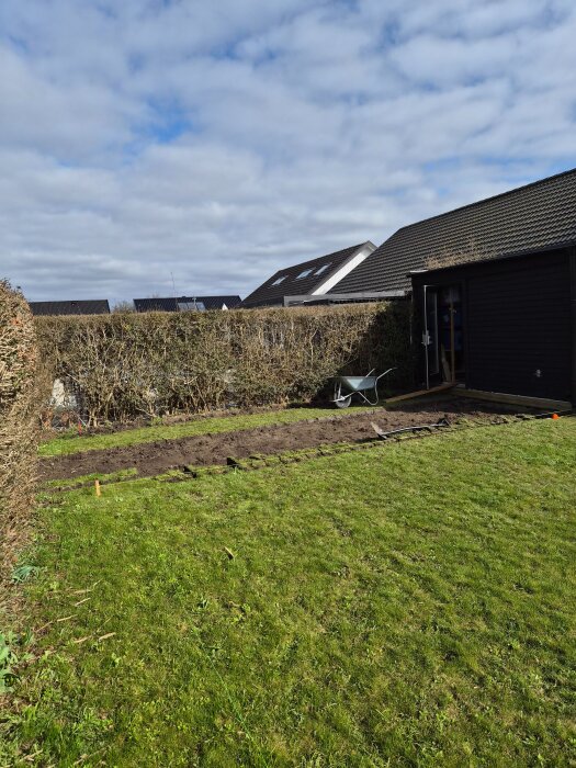 Grävd del av trädgård med gräs, staketlinjer och skottkärra. Förberedd yta för nytt projekt med spabad och trädäck.