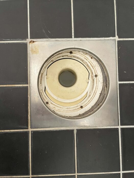 Närbild av en golvbrunn utan vattenlås, omgiven av mörkgrå kakelplattor, med synliga metall- och plastdetaljer.