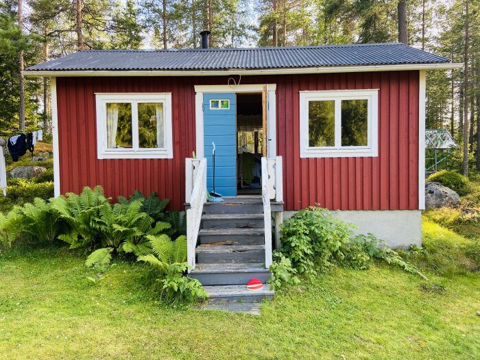 Röd sovstuga med blå dörr, vitmålade fönster och trätrappa på grönskande tomt omgiven av träd.