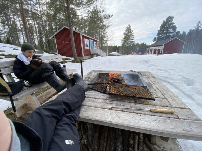 Personer sitter vid en eldplats utomhus med snö runt omkring och läser, med röda stugor i bakgrunden.