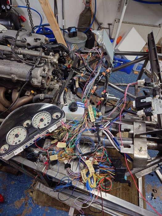 Bild av en bilmotor i ett garage, omgiven av färgglada kablar och instrumentpanel. Arbetet med att färdigställa en tankarmatur pågår.