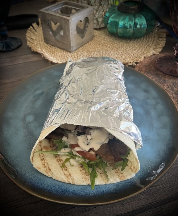 KEBABRULLE med kebabkött, rödlök, ruccola, tomat, gurka och mild kebabdressing i folie på blå tallrik.