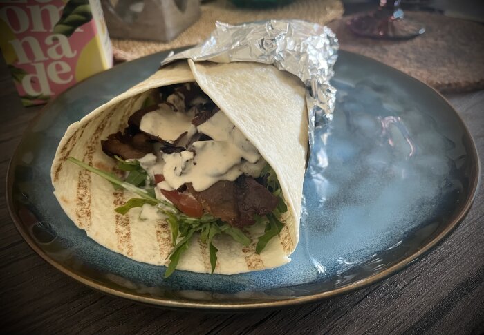 Kebab i tortillabröd med kebabkött, ruccola, rödlök, tomat, gurka och dressing, serverad på tallrik med folietäckt slut.