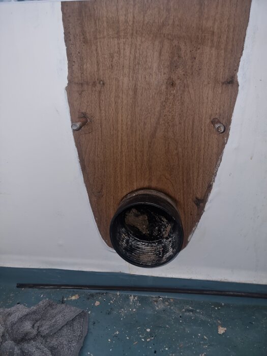 Utsnitt av en båtinteriör med ett runt avgasrör i en träpanel, omgiven av en vit yta. Golvet har smuts och en trasa ligger bredvid.