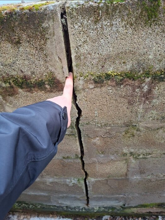 Sprucken mur av betongblock, en hand pekar på spricka. Muren har mossbevuxen yta och sprickan visar tecken på strukturell skada.