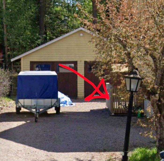 Ingång till källare markerad med röd pil, stor gul byggnad i bakgrunden, grusplan med släpvagn i förgrunden, träd och lampa syns.