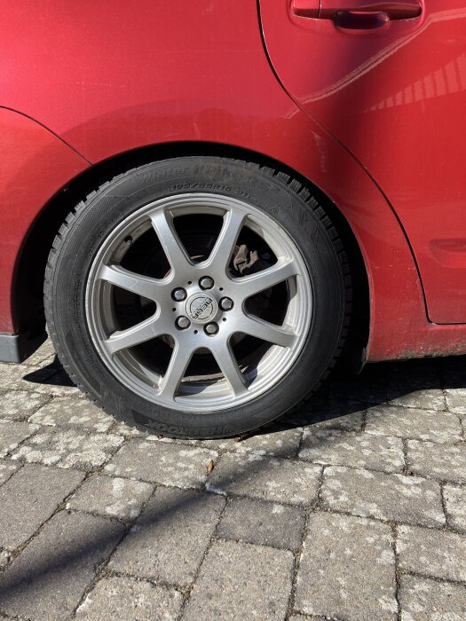 Rött bilkarossutsnitt med ett bakhjul på en Toyota Prius, 195/55R16 vinterdäck på en silverfärgad fälg, stående på stenbeläggning.