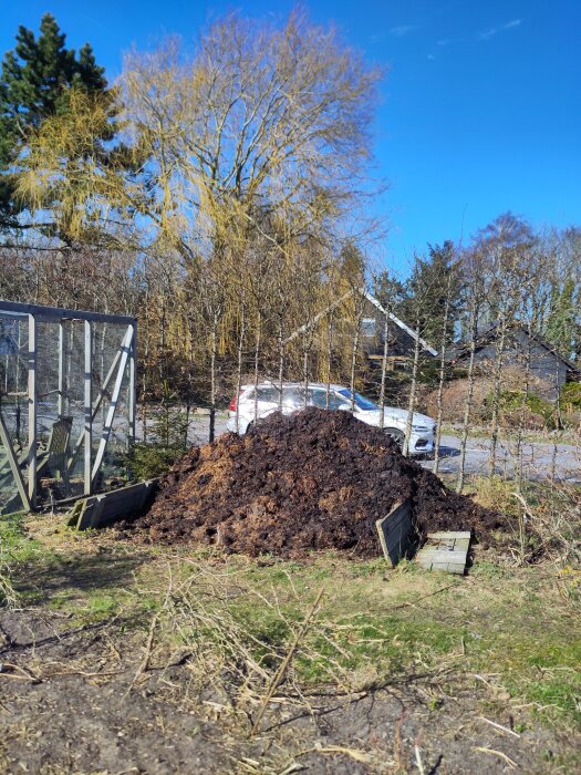 Komposthög i trädgård med kvarvarande snö, omgiven av träd och buskar, under en klarblå himmel.