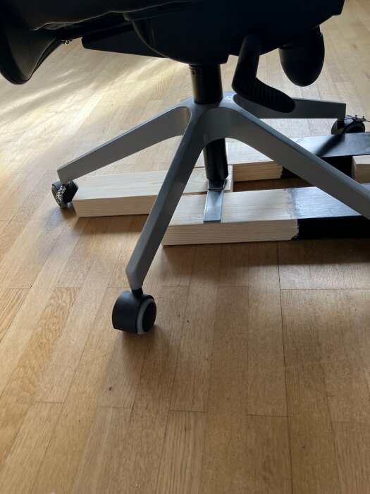 Konstruktion som förhindrar en stol med hjul att välta vid bromsning, placerad på golvet mellan vita träbrädor.