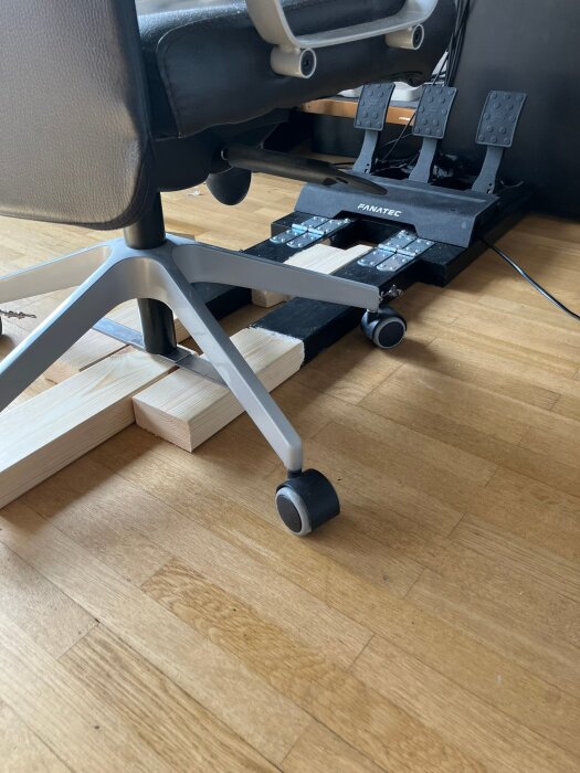 Kontorsstol med hjul på trägolv, placerad på en hemmagjord träkonstruktion för stabilisering. Pedaler i bakgrunden.