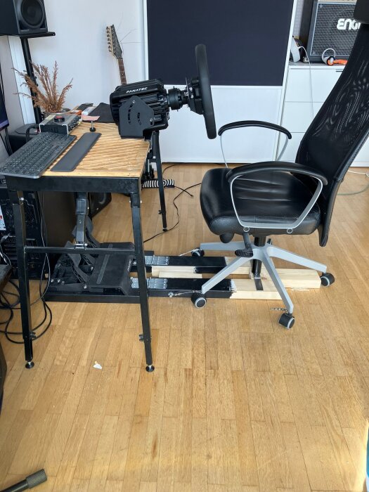 Hemmagjord racing-simulator med ratt monterad på ett skrivbord, kontorsstol på hjul och träkonstruktion för stabilitet på trägolv.