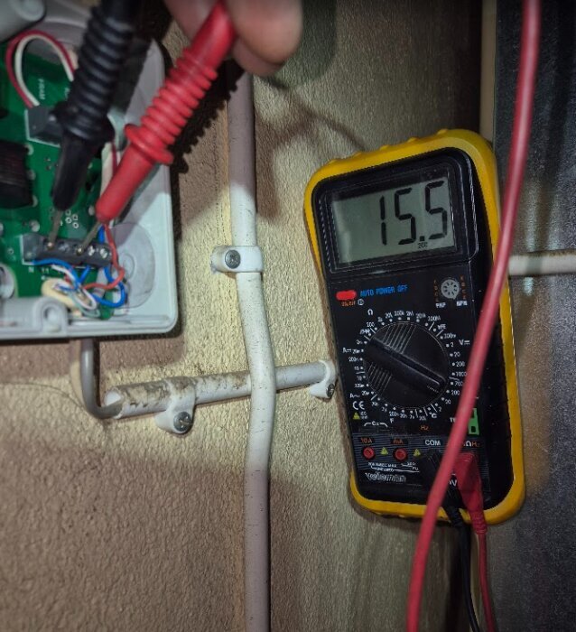Multimeter mäter 15.5 volt i en elinstallation med två prober anslutna till en krets i en öppen kopplingsdosa.