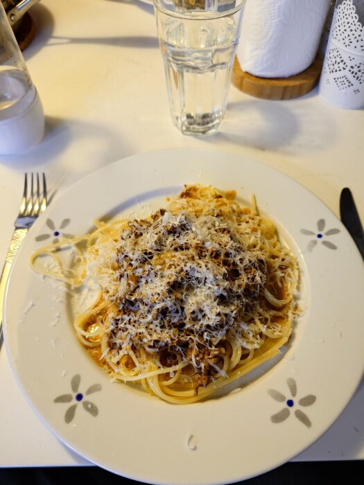 Tallrik med spagetti och köttfärssås toppad med riven ost, bredvid ett glas vatten och bestick på ett bord.