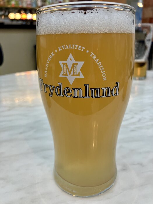 Ett glas fylld med ljusöl på ett marmorerat bord, med texten "Frydenlund" och "Håndverk, Kvalitet, Tradisjonen" tryckt på glaset.