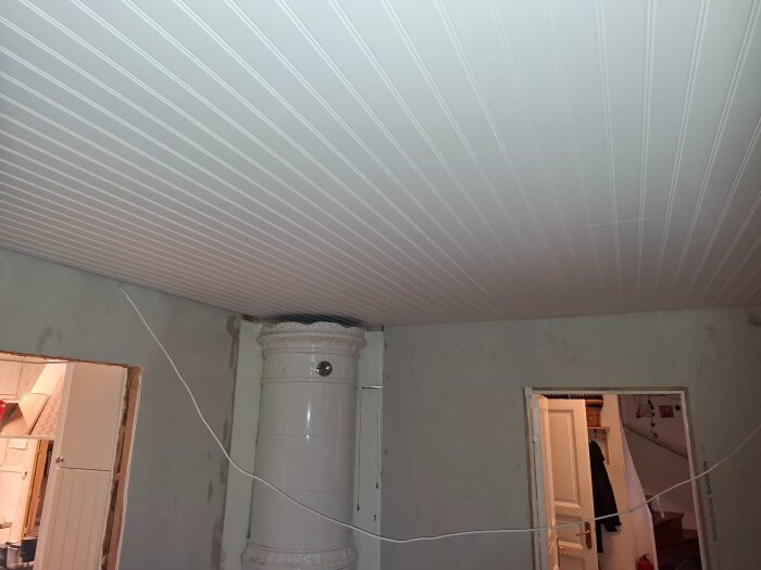Vit träpaneltak med synliga skarvar, dörröppningar, vit kakelugn och hängande kabel i ett rum under renovering.