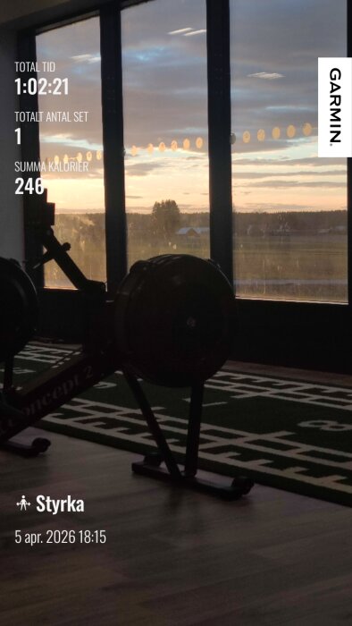 Gymmaskin vid ett fönster med solnedgång i bakgrunden, träningsdata visas på skärmen: tid 1:02:21, antal set 1, kalorier 246, styrketräning.