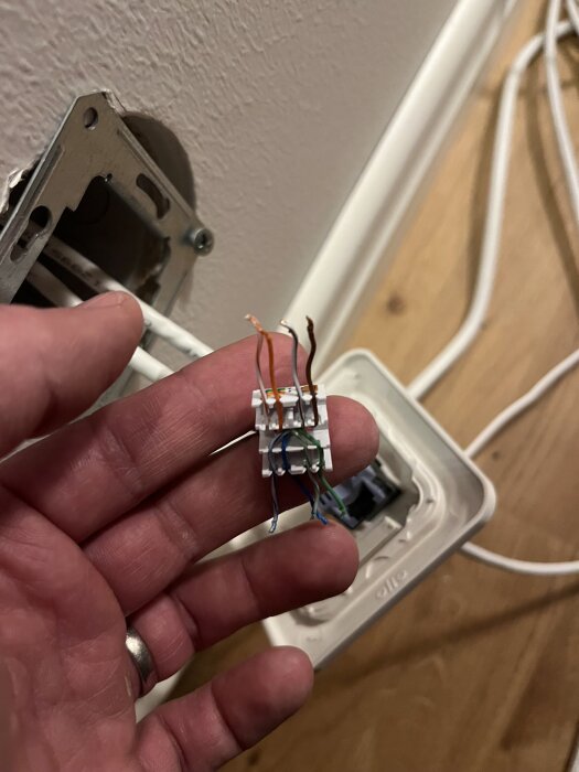 En hand håller en Keystone-kontakt med exponerade kablar, mot en vägg med ett eluttag.