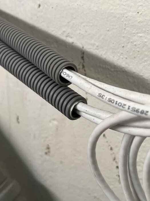 Vita kablar inuti grått flexrör mot en vit vägg, vilket kan indikera installation i ett byggprojekt.