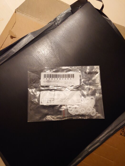 Plastpåse med vita plastbrickor, etikett och streckkod ovanpå svart plastskiva och kartong.