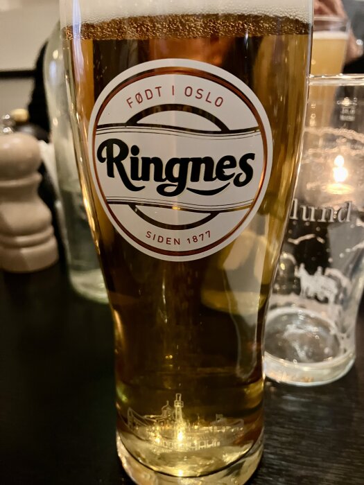 Glas öl med Ringnes-logotypen från Oslo, placerat på ett bord med suddig bakgrund av andra glas och föremål.