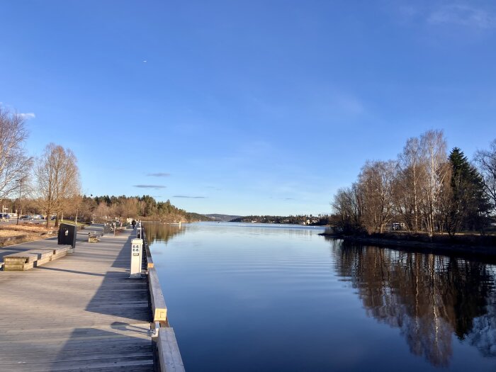 En solig dag vid en brygga med träd längs vattnet och spegelblank yta under en klarblå himmel.