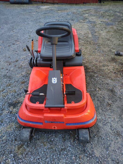 En röd åkgräsklippare av märket Husqvarna Rider 13 står på en grusplan, fotad bakifrån.