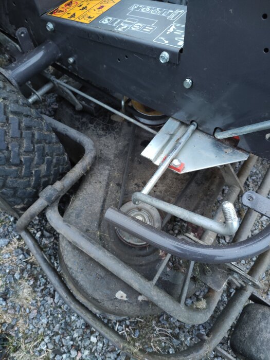 Bild på undersidan av en gräsklippare med motor och hjul synliga, full av smuts och gräsrester.