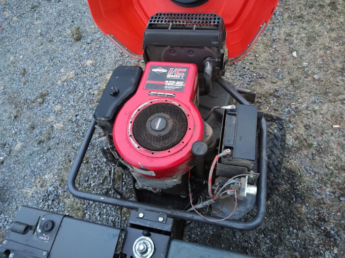 Motor till gräsklippare med Briggs & Stratton 12.5 HP märke, monterad på en ram med stängd huva, ståendes på grusunderlag.