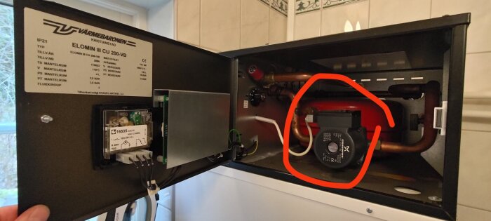 Närbild på en öppen värmepanna med fokus på en cirkulationspump omgiven av rör och elektronik inuti en metallkabinett.