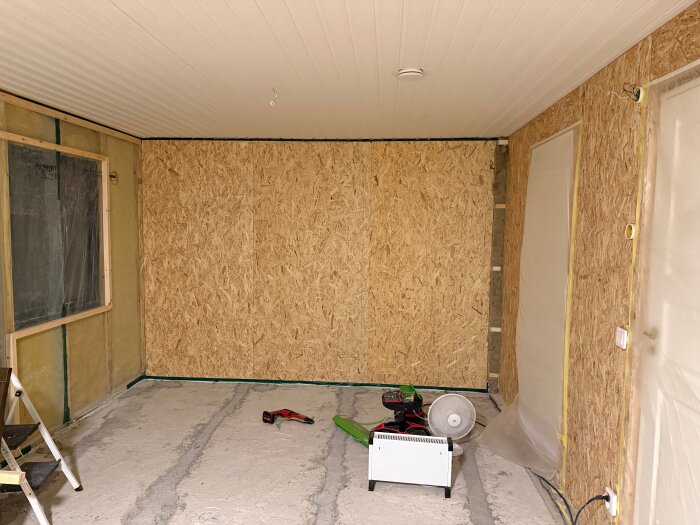Rum under renovering med OSB-skivor på väggarna, verktyg på golvet och en stege vid väggen.