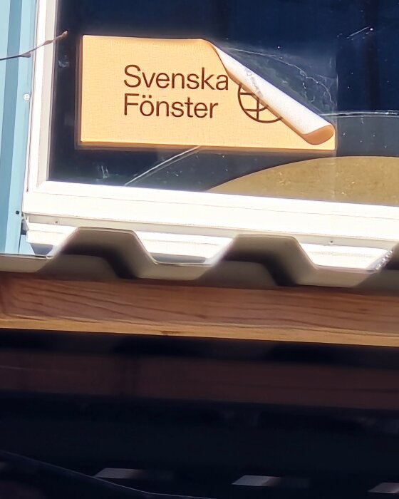 Fönster med etikett som visar texten "Svenska Fönster" och en logotyp, synligt genom en halvöppen del av ett byggnadsmaterial under fönstret.