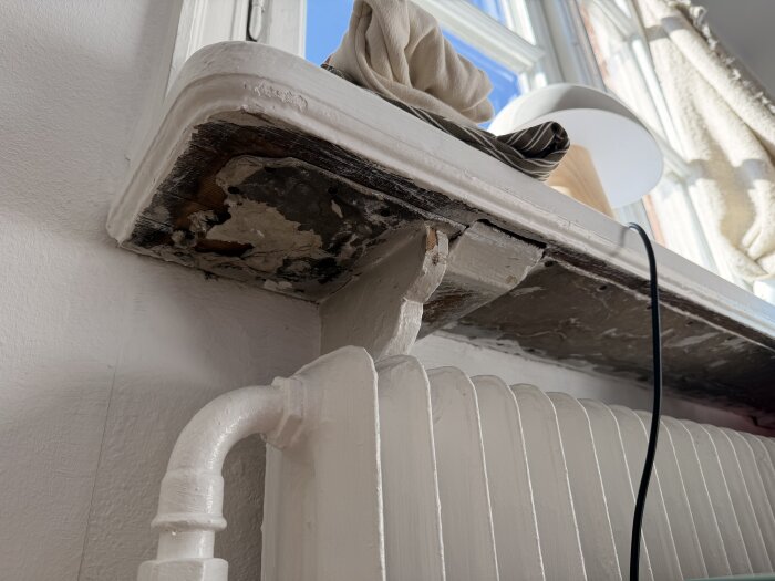 Undersidan av en fönsterbräda med gammalt material, med synliga slitage och ett rör under, i ett hus från tidigt 1900-tal.