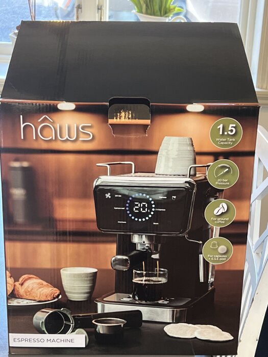 Låda med espressomaskin från Hâws, 1.5L vattentank, 20 bar tryck. Funktionen inkluderar malet kaffe och kapslar, visas med kaffe och croissanter.