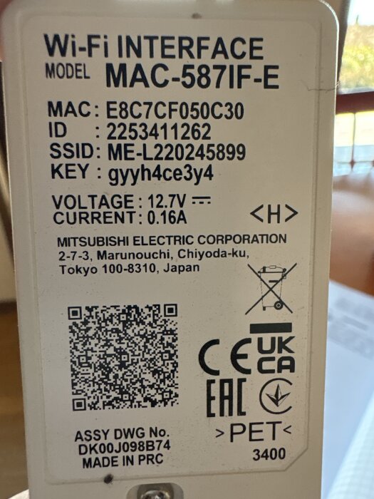 Etikett på en Wi-Fi-interface från Mitsubishi med modellnummer MAC-587IF-E, en MAC-adress, ID, SSID, KEY, spänning och strömstyrka, samt QR-kod och certifieringslogotyper.