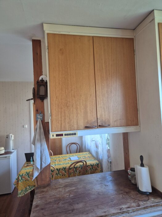 Överskåp i trä med gammal ljusarmatur och två trästolpar. Bakgrund med köksbord täckt av en blommig bordsduk och stolar.