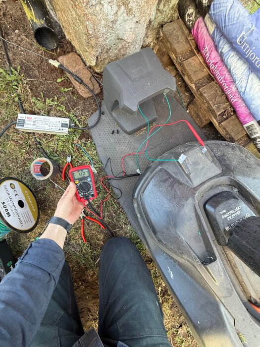 Person justerar elektrisk ström med multimeter och kablar vid laddstation för robotgräsklippare, omgiven av jord och kabelrulle.