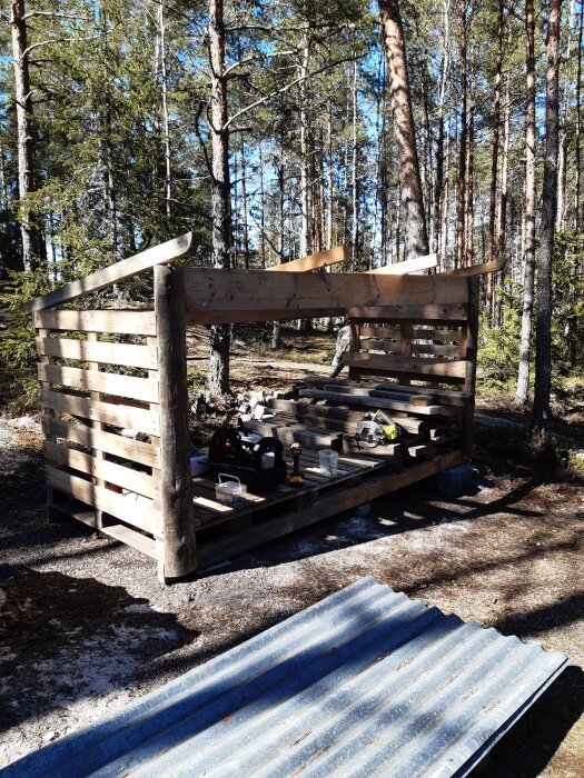 Öppet träskydd under konstruktion i en skog. Byggt av återvunnet trä, med verktyg och trämaterial på plats. Vid sidan syns ett korrugerat metalltak.