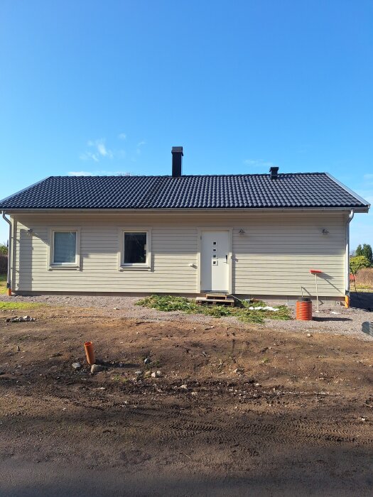 Enkel enplanshus med ljus fasad, svart tak och vita dörrar, omgiven av jord och en grusgång framför entrén, under klarblå himmel.
