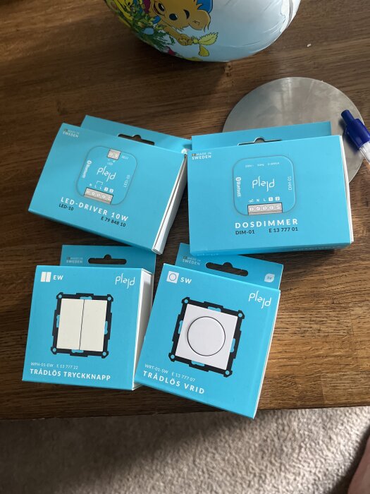 Fyra Plejd förpackningar med trådlös tryckknapp, trådlös vrid, LED-driver och dosdimmer placerade på ett träbord.