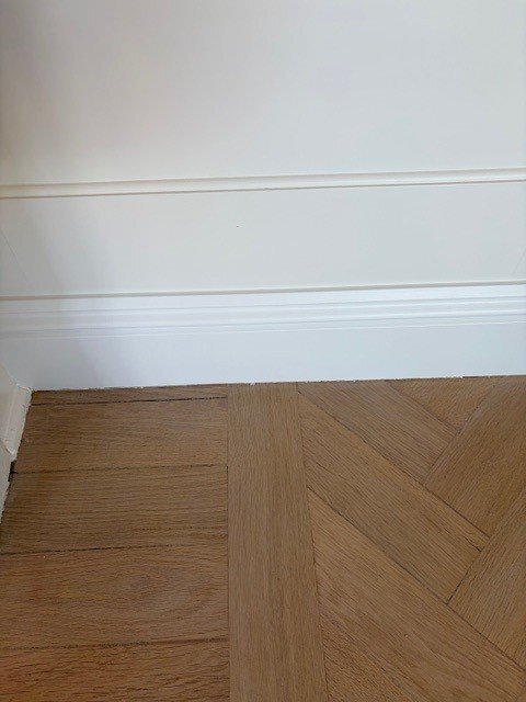 Vit vägg med två lister, den nedersta 1x1 cm, nymålad. Trägolv syns nedanför, med små färgfläckar i hörnet.
