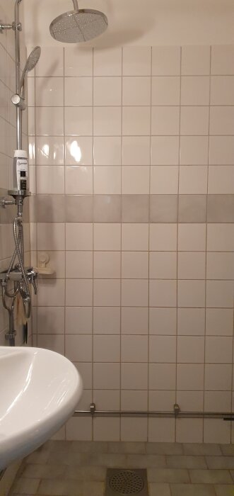 Duschhörna med vitt kakel, en grå rand och duschmunstycke. Fogarna visar missfärgningar, särskilt nära den grå bården.