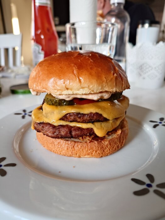 Dubbel cheeseburgare på ett fat, med pickles, sås och smält ost, serverad på ett bord med bakgrund av ketchupflaska och vattenglas.