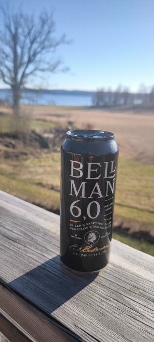Svart ölburk med vit text på ett träräcke med suddig landskapsbakgrund och blå himmel i bakgrunden.