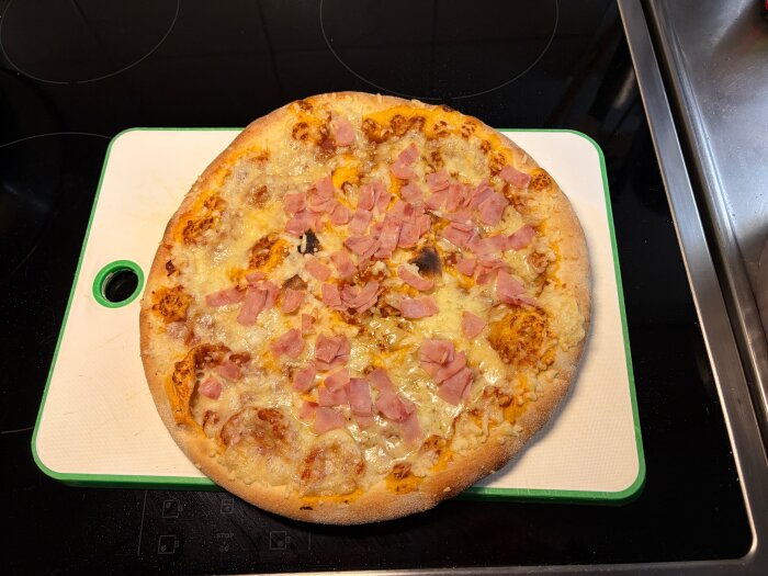 Pizza med ost och skinka på en skärbräda i ett kök.